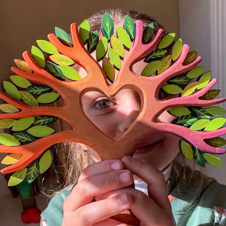 Child peeking through a handcrafted wooden heart tree toy with a bright eye visible through the heart-shaped void