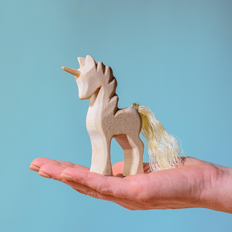 Hand presenting a wooden unicorn figurine, emphasizing the hand-carved details and the contrast of the toy against a soft blue sky background.