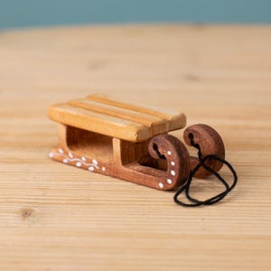 May include: A small wooden sled with a light brown seat and dark brown runners. The runners have white dot and swirl details. A black string is attached to the runners. The sled is on a light brown wooden surface.