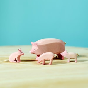 A family of wooden pigs painted in soft pink, with one large pig in the center surrounded by three smaller pigs, on a wooden surface against a turquoise background.