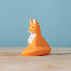Wooden seated fox toy with a detailed white chest, orange body, and black-tipped ears on a wooden surface with a blue backdrop.