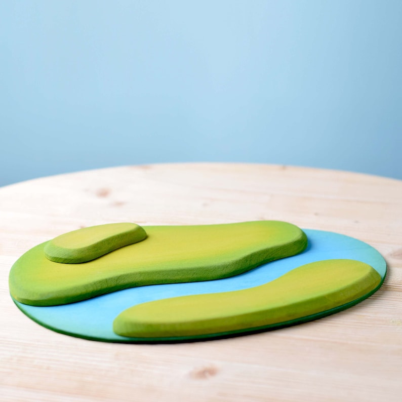 Blank wooden play mat with a river and grass design, crafted for imaginative play, on a wooden surface with a blue wall behind.