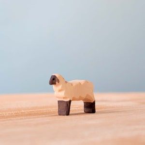 Small wooden sheep toy standing on a wooden surface, with dark facial and leg features, against a light blue background.