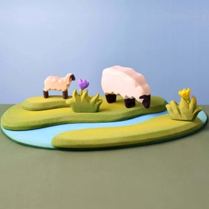 Wooden playset featuring two sheep by a water feature with greenery and purple flower, against a pale blue background.