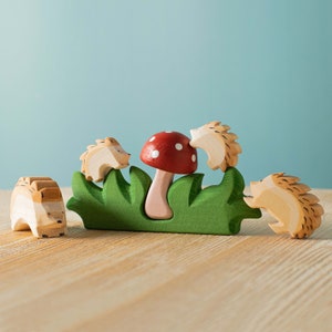 A red mushroom with white spots surrounded by green grass and three wooden hedgehog toys on a light wood surface.