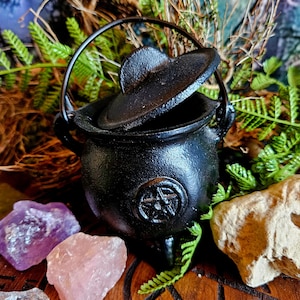Op de afbeelding: Een kleine zwarte gietijzeren ketel met een pentagram-ontwerp en een deksel. De ketel staat op een houten oppervlak met groen gebladerte en kristallen.