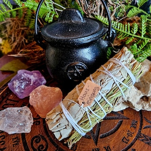 Könnte beinhalten: Ein weißer Salbei-Reinigungsstab mit einem Etikett mit der Aufschrift "Haus - reinigung" ist mit weißem Garn gebunden und auf einer Holzoberfläche mit einem schwarzen Kessel, rosa und violetten Kristallen und einem Stück weißem Quarz platziert.