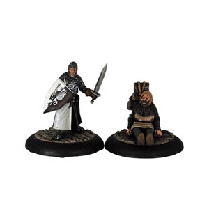 Pode incluir: Duas figuras em miniatura retratando figuras medievais. Uma figura é um cavaleiro em armadura de malha, segurando uma espada e um escudo. A outra figura é uma pessoa sentada em uma túnica com capuz. Ambas as figuras estão em bases circulares.