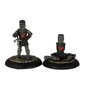 Könnte beinhalten: Zwei Miniaturfiguren von Rittern in schwarzer Rüstung mit roten Kreuzen auf der Brust. Ein Ritter steht mit einem Schwert, während der andere mit seinem zerbrochenen Schwert am Boden liegt.