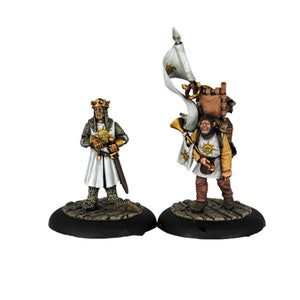 Könnte beinhalten: Zwei bemalte Miniaturfiguren, eine ein Ritter in weißer Rüstung mit Krone und Schwert, die andere eine Figur in einer weißen Tunika, die eine Fahne und ein Horn trägt. Beide Figuren haben ein Sonnensymbol auf der Brust.