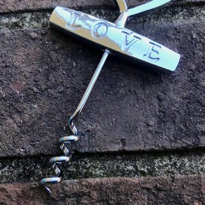 May include: A silver corkscrew with a handle that says "LOVE" on it. The corkscrew is lying on a brick surface.