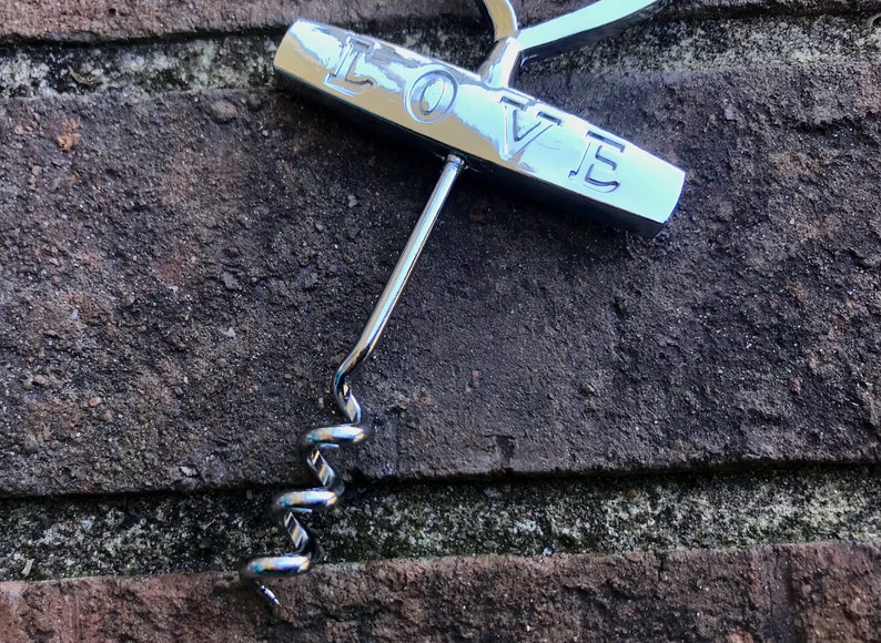 May include: A silver corkscrew with a handle that says "LOVE" on it. The corkscrew is lying on a brick surface.