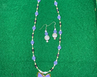 Puede incluir: Un collar con un colgante de mariposa morado, una cadena de cuentas y pendientes a juego. Las cuentas son una mezcla de azul claro, blanco y negro.