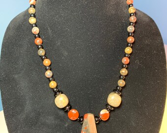 Puede incluir: Un collar con un colgante de piedra grande y multicolor. El collar está hecho de cuentas negras, marrones y beige.