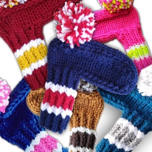 May include: A collection of colorful knitted golf club head covers with pom-poms. The covers are in various colors, including red, blue, pink, brown, and gray. Each cover has a white stripe and a pom-pom on top.