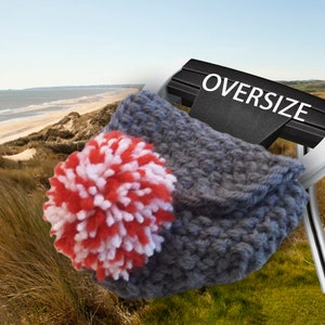 May include: A gray, oversized crocheted hat with a large red and white pom-pom. The word "OVERSIZE" is displayed on a black sign. The background features a beach scene with sand dunes and ocean.