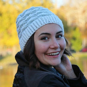 Peut inclure: Une femme portant un bonnet en tricot blanc et gris avec un motif en tricot torsadé. Elle sourit et regarde de côté.