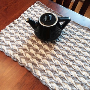 May include: A gray and white crocheted placemat with a black teapot on top.