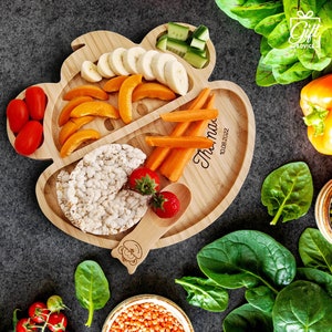May include: A wooden divided plate shaped like a bear with a spoon. The plate has sections for rice cakes, fruit, vegetables, and a small spoon. The plate is personalized with the name "Theo" and the date "10.08.2022".
