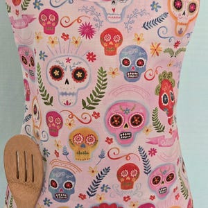 May include: A pink apron with a colorful Day of the Dead sugar skull pattern. The apron has a light blue neck strap and a wooden spoon in the front pocket. The design includes flowers and decorative elements.