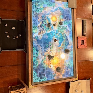 May include: A wooden-framed tabletop gaming display with a colorful map, dice trays, and accessories. The display shows a grid-based map with miniature figures. Additional items include a remote control and a small wooden box.