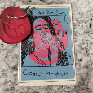 May include: A blue and pink illustrated card with a woman singing into a microphone. The text on the card reads "Didi Bidi Bom Bom" and "Como me duele".