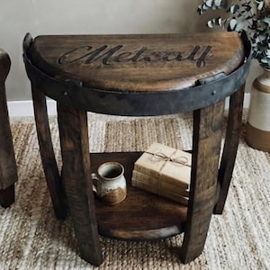 May include: A semi-circular wooden side table crafted from a barrel, featuring a dark brown finish and a metal band around the edge. The top surface is engraved with the name Metcalf in script. A lower shelf holds a ceramic mug and three books.