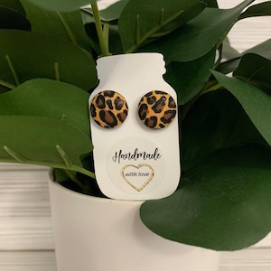 May include: Two round stud earrings with a leopard print pattern. The earrings are displayed on a white card with the text "Handmade with love" and a gold heart. The background features green leaves and a white pot.