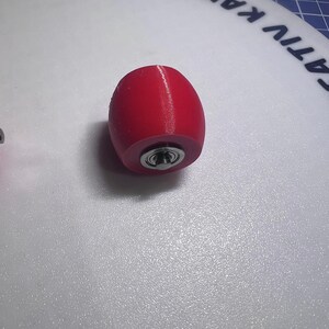 Puede incluir: Un objeto de color rojo brillante con forma de barril y acabado mate liso. Un rodamiento de bolas met&aacute;lico circular est&aacute; incrustado en el centro de la cara frontal, con un anillo exterior plateado y un peque&ntilde;o punto central oscuro.