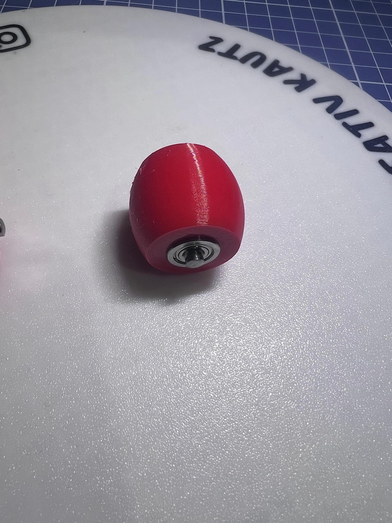 Puede incluir: Un objeto de color rojo brillante con forma de barril y acabado mate liso. Un rodamiento de bolas met&aacute;lico circular est&aacute; incrustado en el centro de la cara frontal, con un anillo exterior plateado y un peque&ntilde;o punto central oscuro.