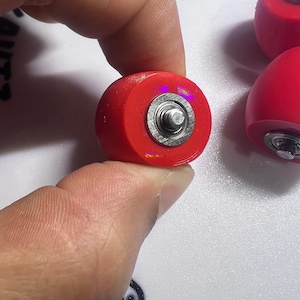 Puede incluir: Un conjunto de ruedas circulares para patines de color rojo brillante, cada una con un rodamiento de bolas central de metal plateado. Las ruedas tienen una superficie exterior roja lisa y un componente central met&aacute;lico visible en el centro.