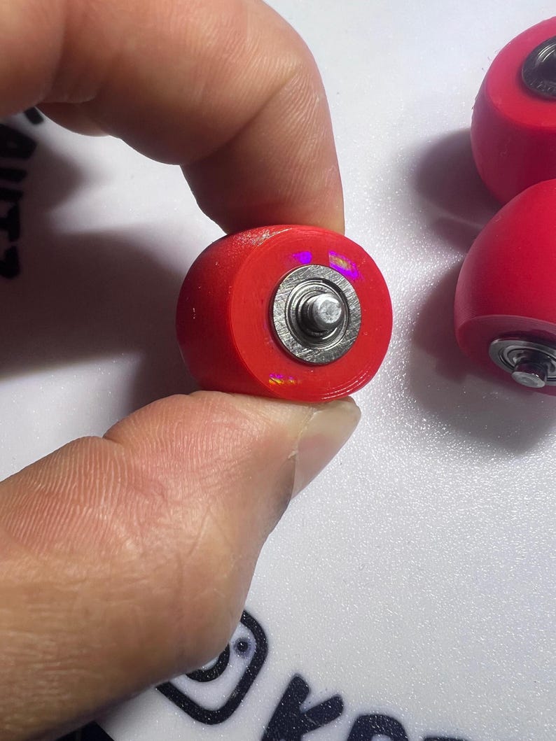 Puede incluir: Un conjunto de ruedas circulares para patines de color rojo brillante, cada una con un rodamiento de bolas central de metal plateado. Las ruedas tienen una superficie exterior roja lisa y un componente central met&aacute;lico visible en el centro.