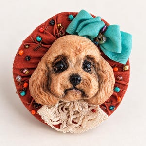 Puede incluir: Un broche redondo con la cara de un perro color canela esculpida, con ojos y nariz oscuros, sobre un fondo de tela color óxido. La pieza está decorada con un lazo de cinta turquesa, pequeñas cuentas de colores y un cuello de encaje blanco.