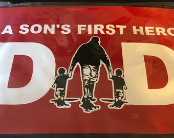 以下が含まれることがあります： 白と赤の看板に、白い文字で「A Son's First Hero DAD」と書かれている。看板の中央には、父親と2人の子供が歩く黒いシルエットが描かれている。