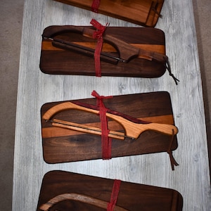 May include: Four wooden cheese slicers with red ribbon accents are displayed on a light gray surface. Each slicer has a wooden handle and a wire blade, and is attached to a rectangular wooden cutting board. The wood tones vary from light to dark.