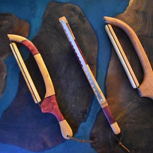May include: Three wooden kitchen tools with curved and straight blades. The tools feature natural wood tones, with accents of red and purple. Each tool has a leather cord attached to the handle.