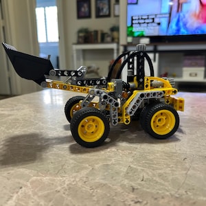 May include: A construction vehicle model built from yellow, black, and gray plastic building blocks. The vehicle features a large black front bucket, a roll cage structure, and four yellow wheels with black tires, showcasing intricate mechanical joints.