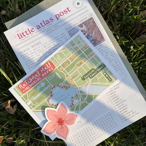 以下が含まれることがあります： 「little atlas post」というタイトルの印刷されたニュースレター。ナショナルモールと記念公園の地図、ピンクの花のステッカー、単語検索があります。ニュースレターは緑の草の上にあります。