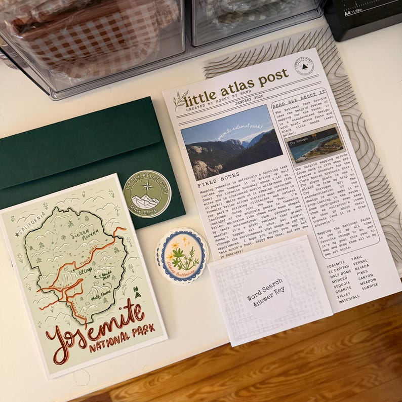 May include: A collection of stationery items featuring a Yosemite National Park theme. Includes a green envelope with a circular sticker, a map-style postcard, a newspaper-style print, and a floral sticker. The postcard shows a map of Yosemite.