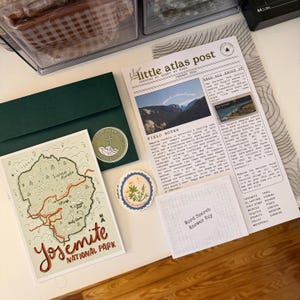 May include: A selection of stationery items, including a green envelope, a map of Yosemite National Park, a newspaper-style print, and stickers. The map features a detailed illustration with red and green accents. The newspaper has text and images.