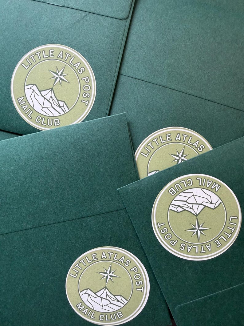 May include: A collection of dark green envelopes with circular stickers. The stickers feature the text "LITTLE ATLAS POST MAIL CLUB" around a mountain and compass rose design in white and olive green.