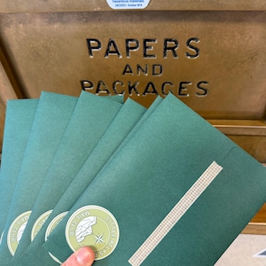May include: A hand holding a stack of green envelopes with a white and green circular sticker that reads "Little Atlas Mail Club". The envelopes are sealed with a piece of tape. The background shows a USPS mailbox with the words "Papers and Packages".
