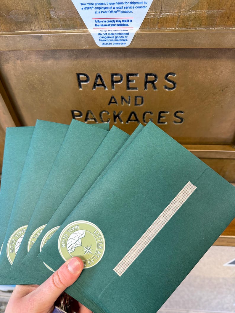 May include: A hand holding a stack of green envelopes with a white and green circular sticker that reads "Little Atlas Mail Club". The envelopes are sealed with a piece of tape. The background shows a USPS mailbox with the words "Papers and Packages".