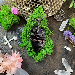 May include: A black obsidian pendant wrapped in copper wire, hanging from a brown leather cord. The pendant is resting on a bed of green moss.