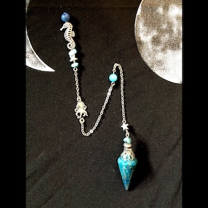Könnte beinhalten: Ein silbernes Pendel mit einem blauen und türkisfarbenen Stein, verziert mit Seepferdchen-, Oktopus- und Stern-Anhängern. Das Pendel hängt an einer silbernen Kette mit blauen Perlen. Das Design hat ein himmlisches und nautisches Thema.