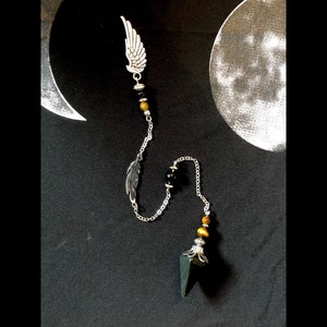 May include: A black and gold pendulum with a silver chain and two feather charms. The pendulum is made of a black stone with a pointed tip and a silver cap. The chain is adorned with black and gold beads.