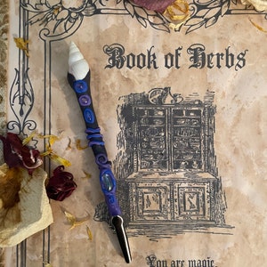 May include: A purple and blue wand with a seashell top and a black handle. The wand is lying on a vintage book with the text "Book of Herbs" and the quote "You are magic. Let your intuition lead the way."