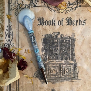 May include: A blue and white seashell-shaped wand with a seahorse charm and blue beads. The wand is resting on a vintage book with the text "Book of Herbs" and the quote "You are magic. Let your intuition lead the way."