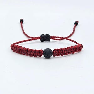 Puede incluir: Un brazalete trenzado rojo con una cuenta de lava negra. El brazalete tiene lazos ajustables en cada extremo.