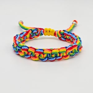 Puede incluir: Un brazalete trenzado de colores con hilos rojos, naranjas, amarillos, verdes, azules y azul claro. El brazalete está atado con un nudo y tiene cuentas de metal plateadas en los extremos.